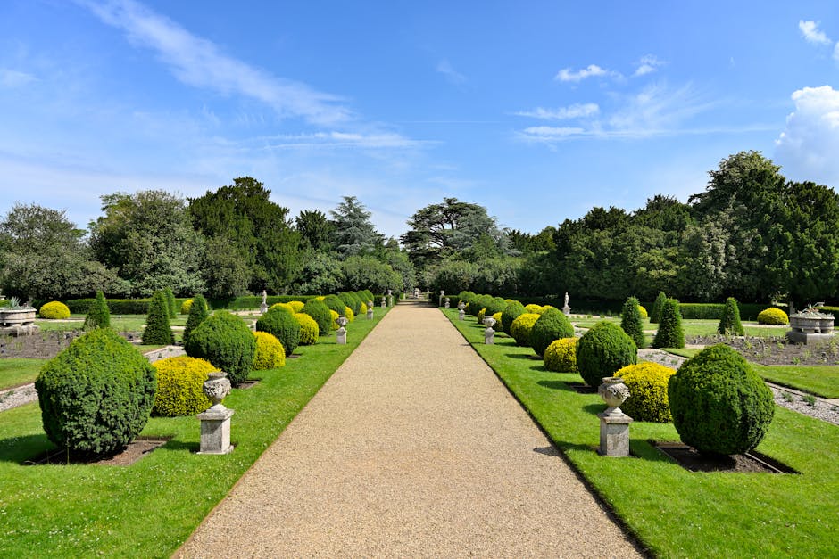 A symmetrical garden pathway in a formal landscape setting, with a wide, gravel-lined walkway extending straight into the distance, flanked by meticulously trimmed round and conical shrubs with lush g