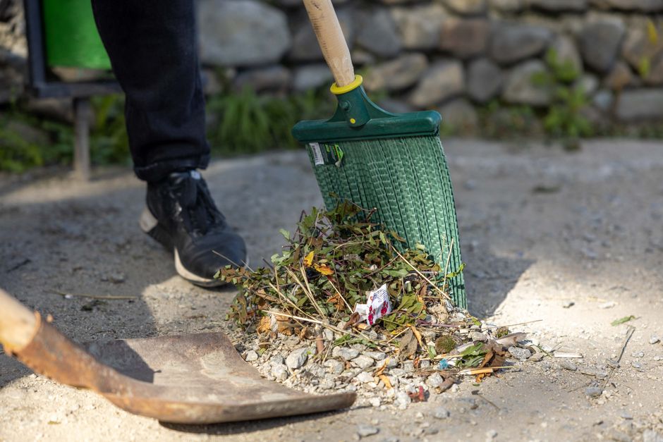 A person wearing black pants and black sneakers is using a green hand broom with a wooden handle to sweep dry leaves, twigs, and small debris into a pile on a gravel pathway. The pathway has a section