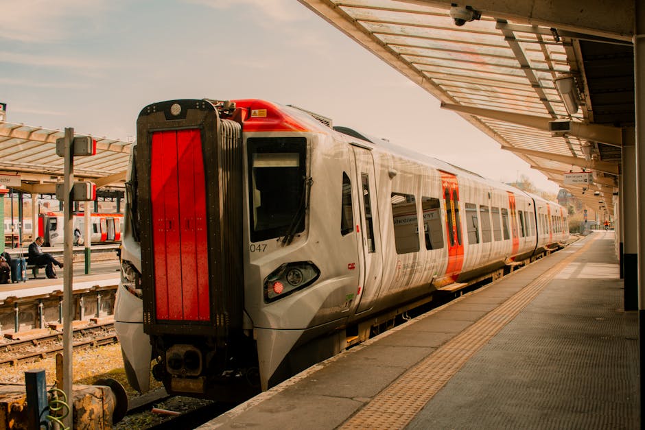 A modern commuter train with a sleek, aerodynamic design is seen at a train station platform, partially covered by a metal and glass canopy. The train has a white exterior with bold red accents along